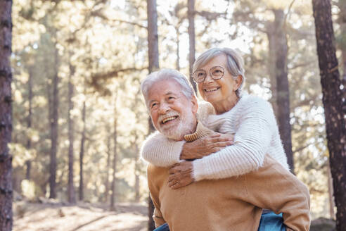 Glücklicher älterer Mann, der eine Frau mit Brille im Wald huckepack nimmt - SIPF02782