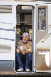 Smiling senior man with coffee cup sitting at door of motor home - SIPF02783