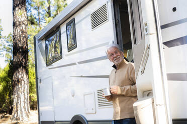 Smiling senior man with coffee cup standing at door of motor home - SIPF02784