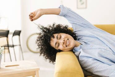 Smiling young woman with curly hair lying on sofa at home - XLGF02813