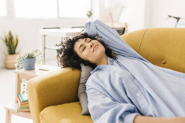 Smiling young woman with eyes closed resting on sofa at home - XLGF02814