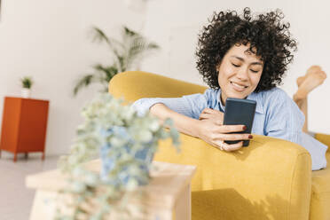 Smiling young woman using smart phone lying on sofa at home - XLGF02816