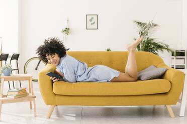 Happy young woman surfing net through smart phone lying on sofa in living room at home - XLGF02818