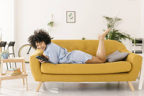 Glückliche junge Frau, die auf dem Sofa im Wohnzimmer zu Hause liegt und mit ihrem Smartphone im Internet surft - XLGF02818