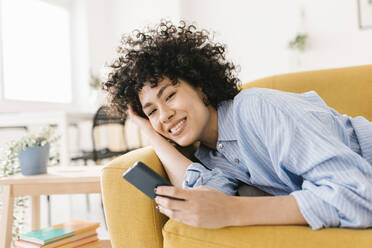 Happy woman holding mobile phone lying on sofa in living room - XLGF02821