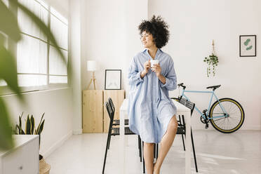 Smiling young woman leaning on table with coffee cup at home - XLGF02825