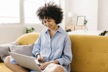 Smiling young woman using laptop sitting on sofa in living room - XLGF02836