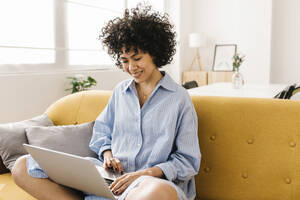Lächelnde junge Frau mit Laptop auf dem Sofa im Wohnzimmer sitzend - XLGF02836