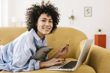 Glückliche junge Frau mit Kreditkarte und Laptop auf dem Sofa zu Hause liegend - XLGF02840