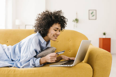 Smiling woman holding credit card using laptop lying on sofa in living room at home - XLGF02841