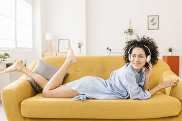 Smiling young woman wearing wireless headphones lying on sofa at home - XLGF02844