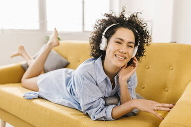 Smiling woman with headphones lying on sofa in living room at home - XLGF02845