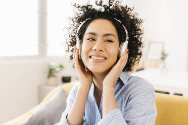 Happy woman with curly hair listening music through wireless headphones at home - XLGF02847