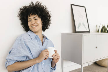 Glückliche junge Frau mit Kaffeetasse, die zu Hause an einer weißen Wand sitzt - XLGF02851
