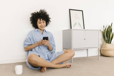 Happy young woman using smart phone sitting cross-legged at home - XLGF02853