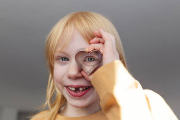 Happy blond girl holding heart shape cookie cutter in front of eye at home - IHF00766