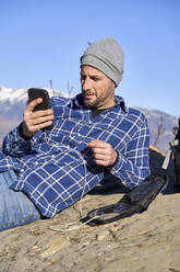 Man wearing knit hat surfing net through smart phone lying by backpack on sunny day - VEGF05326