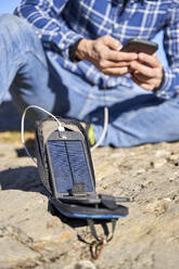 Mann, der an einem sonnigen Tag sein Mobiltelefon mit einem tragbaren Solarmodul auflädt - VEGF05329