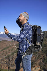 Man wearing backpack holding smart phone charged through portable solar panel on sunny day - VEGF05330