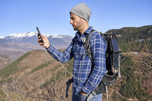 Mann steht mit Hand in der Tasche und überprüft sein Smartphone an einem sonnigen Tag - VEGF05331