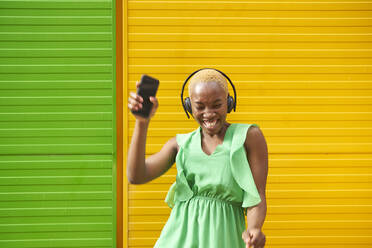 Cheerful woman with eyes closed holding smart phone dancing in front of green and yellow wall - VEGF05343