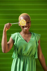 Glückliche Frau, die ihr Auge mit einem gelben Lutscher bedeckt, steht vor einer grünen Wand - VEGF05344