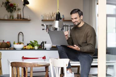 Smiling young man using laptop siting on table in kitchen at home - PESF03438