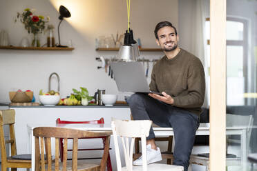 Smiling freelancer with laptop sitting on table in kitchen at home - PESF03439