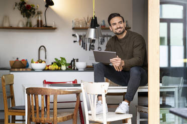 Smiling young man with tablet PC sitting on table in kitchen at home - PESF03441