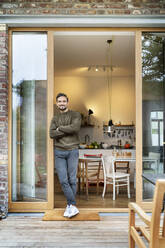 Smiling young man standing with arms crossed leaning on doorway at patio - PESF03444