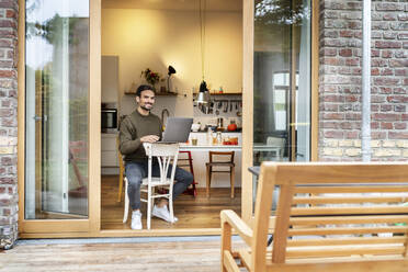 Smiling young man with laptop sitting on chair in kitchen - PESF03446