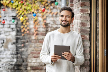 Lächelnder junger Mann mit Tablet-PC an einer Backsteinmauer - PESF03449
