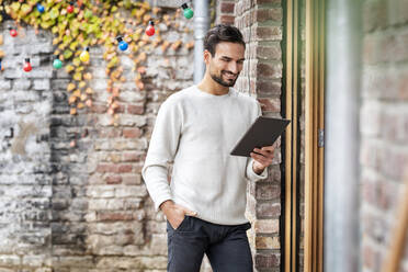 Happy freelancer with hand in pocket using tablet PC by brick wall - PESF03452