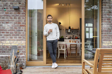 Young man holding glass of coffee standing with hand in pocket leaning on doorway - PESF03454