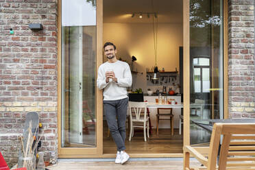 Smiling young man holding glass of coffee leaning on doorway - PESF03455