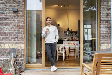 Smiling man with glass of coffee leaning on doorway - PESF03457