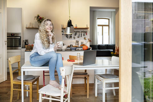 Glückliche junge Frau mit Kaffeetasse und Hand am Kinn in der Küche sitzend - PESF03463