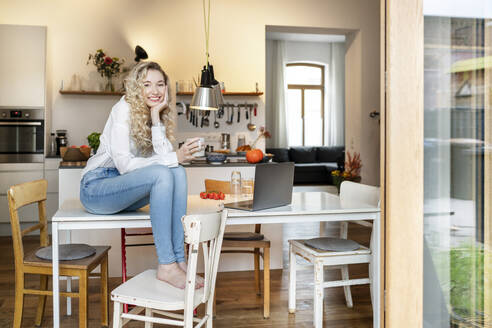 Glückliche blonde Frau mit Kaffeetasse, die mit dem Kopf in der Hand auf einem Tisch sitzt - PESF03464