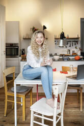 Glückliche schöne Frau mit blondem Haar, die eine Kaffeetasse hält und auf einem Tisch in der Küche sitzt - PESF03465