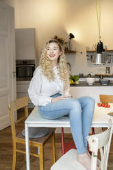Cheerful blond woman holding coffee cup sitting on table in kitchen at home - PESF03466