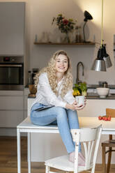 Smiling blond woman holding coffee cup sitting with laptop on table in kitchen at home - PESF03471