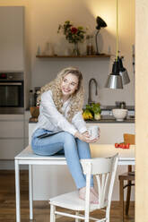 Smiling blond freelancer holding coffee cup sitting with laptop on table in kitchen at home - PESF03472