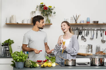 Young man talking with happy girlfriend holding pasta standing in kitchen at home - PESF03475
