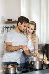 Lächelnde blonde Frau sieht ihren Freund beim Nudelkochen in der Küche an - PESF03483