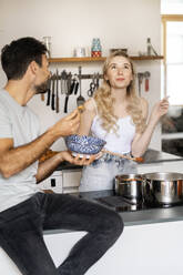 Young man looking at girlfriend tasting food in kitchen - PESF03487