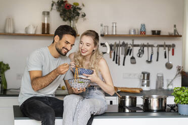 Smiling young woman looking at man eating spaghetti sitting on kitchen island at home - PESF03491