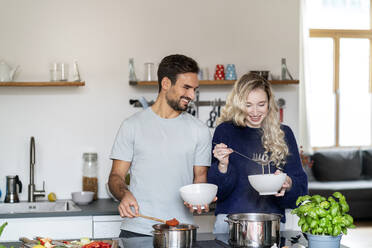 Glückliches Paar serviert Essen in Schüsseln an der Kücheninsel stehend - PESF03494