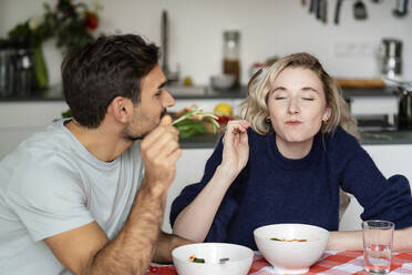 Junger Mann sieht seine Freundin an, die mit geschlossenen Augen am Tisch sitzend isst - PESF03499