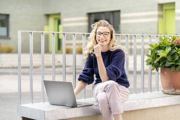 Lächelnder blonder Freiberufler mit Brille sitzt mit Laptop an der Wand - PESF03510