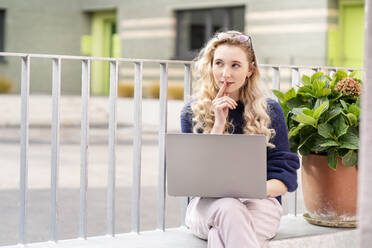 Thoughtful young woman with laptop sitting on wall leger - PESF03513